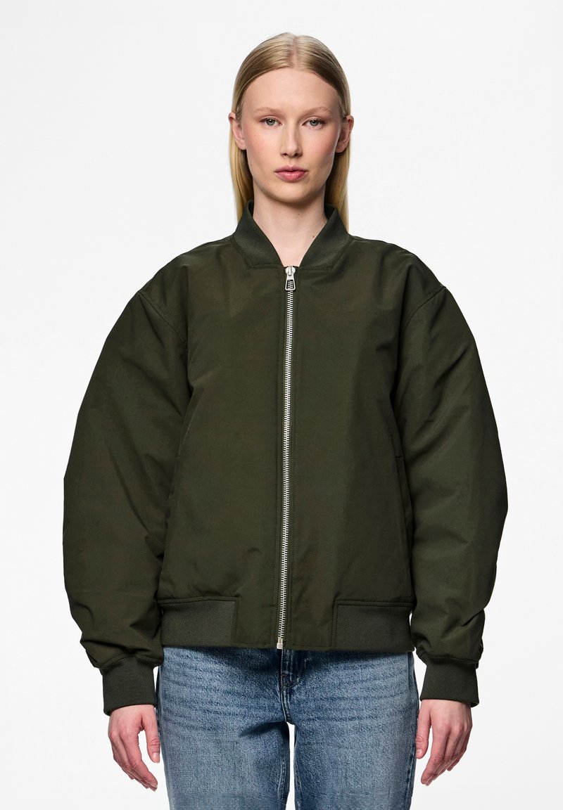 Casaco bomber verde-azeitona com fecho de correr na frente, punhos canelados e corte oversized. Apresenta dois bolsos laterais. Usado sobre um denim casual.