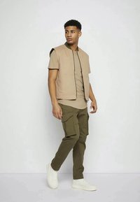 Casaco sem mangas beige sobre uma camisa castanha, combinado com calças cargo verdes-oliva. Apresenta dois bolsos frontais e um design de corte descontraído, usado com ténis brancos.