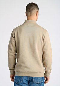 Sudadera beige con cuello alto y mangas largas, con un acabado texturizado. Combinada con jeans de mezclilla azul.
