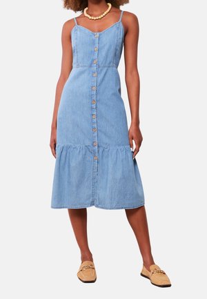 Kvinde iført en lyseblå ærmeløs jeans midi-kjole med knapper foran og flæsekant, beige loafers og en træperlehalskæde.