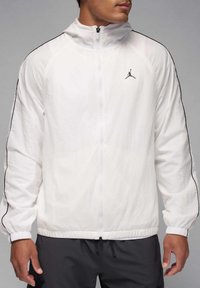 Chaqueta cortavientos blanca con rayas laterales negras, cierre de cremallera, cuello alto y un pequeño logo negro en el pecho. Tejido texturizado.