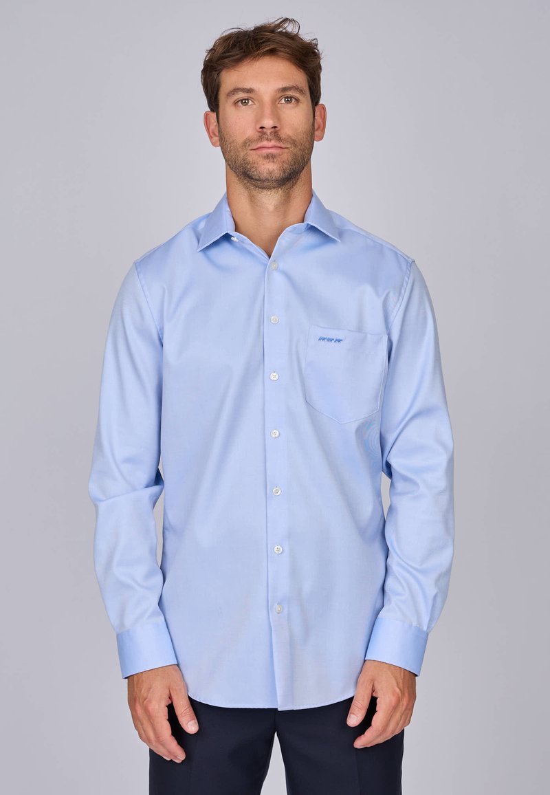 Chemise bleu clair à manches longues, avec un col structuré, une poche poitrine et un logo discret brodé en bleu sur la poche.
