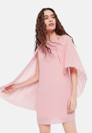 Giovane donna con lunghi capelli ricci che indossa un vestito rosa chiaro con dettaglio mantella plissettata e un bottone sulla spalla.
