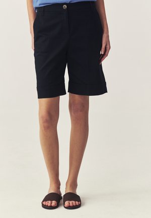Kvinde iført sorte knælange, skræddersyede shorts med opsmøgede kanter og brune slip-in sandaler, stående mod en ensfarvet baggrund.
