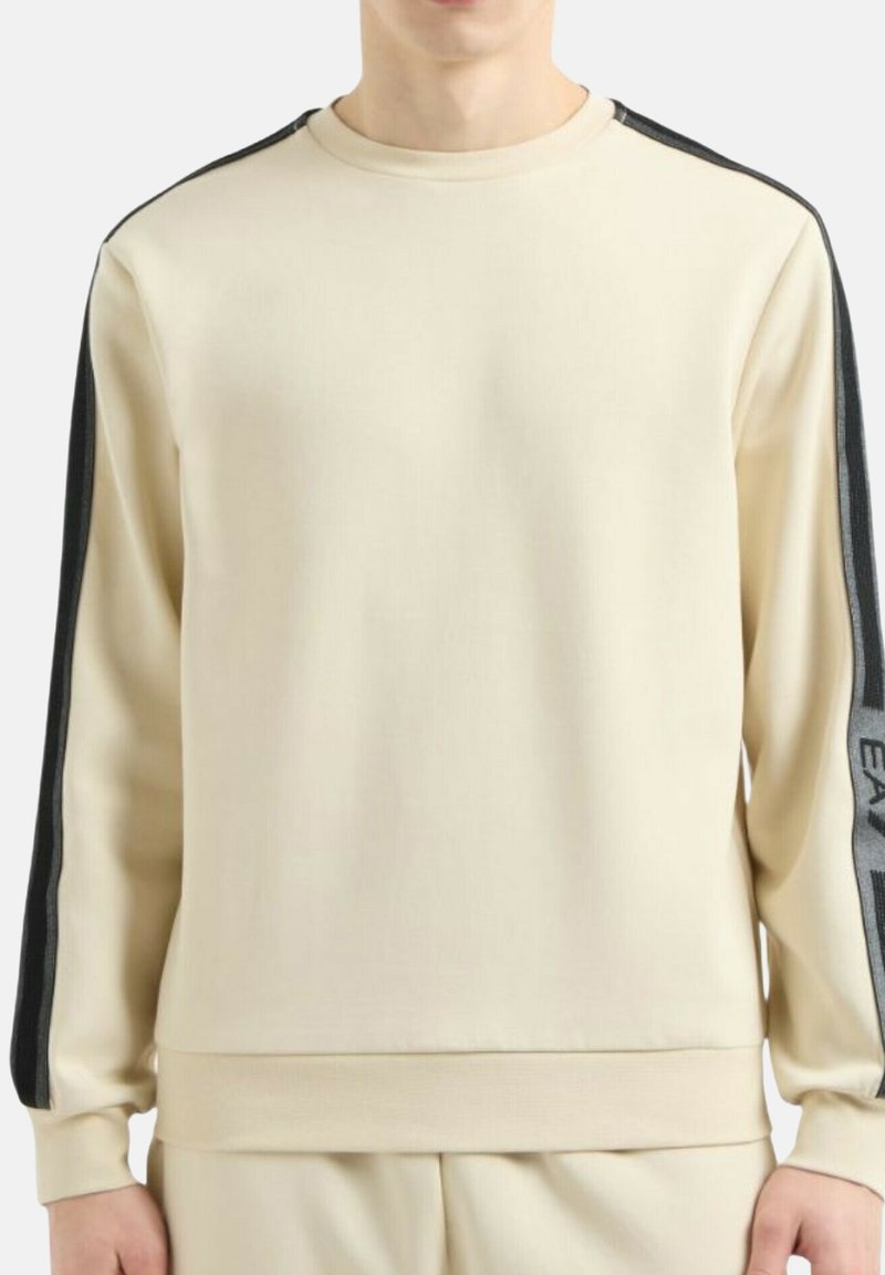 Uomo che indossa una felpa beige a maniche lunghe con strisce nere e testo sulle maniche, abbinata a pantaloni beige coordinati.