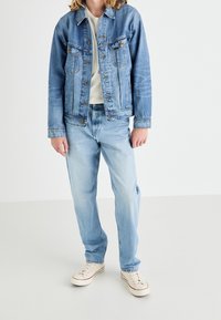 Ljusblå denimjacka med två bröstfickor, tillsammans med ljusblå jeans och beige sneakers. Enkel design, avslappnad passform.
