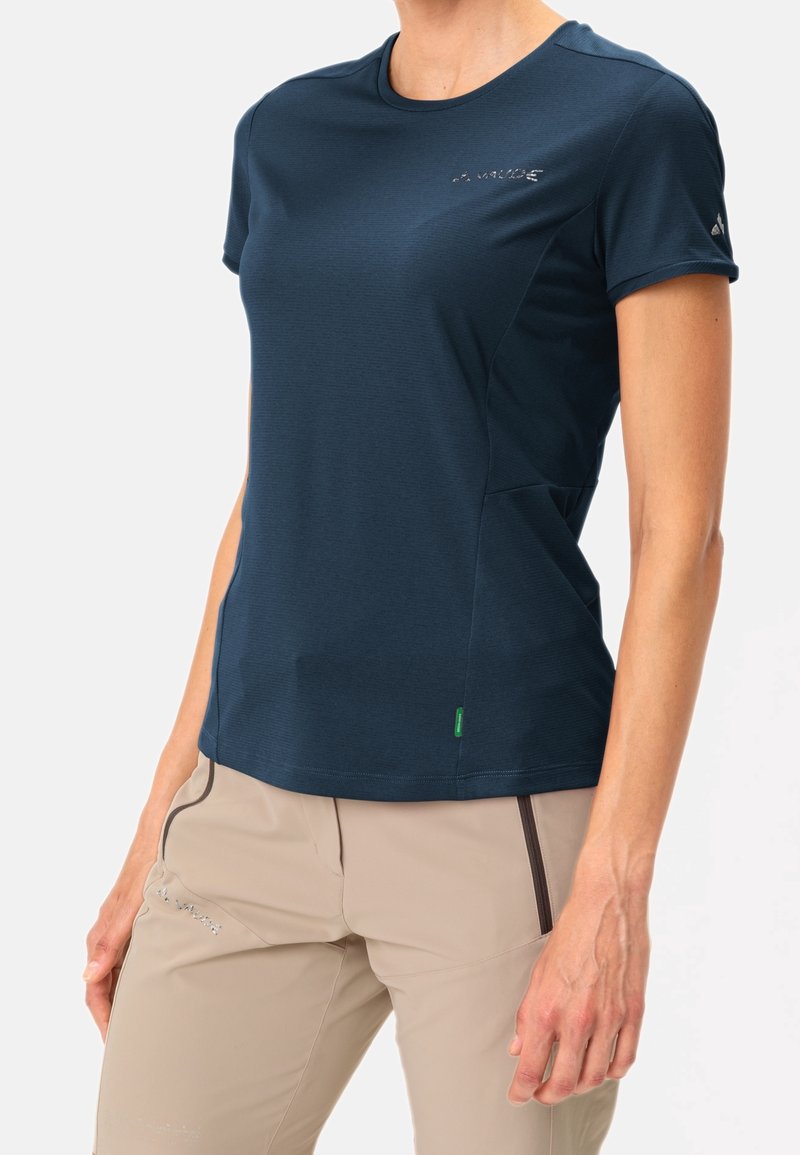 Femme portant un t-shirt bleu foncé ajusté à manches courtes et un pantalon beige avec des détails de fermeture éclair, debout devant un fond clair uni.