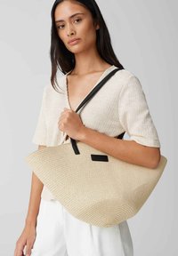 Borsa a spalla intrecciata beige con una forma curvata, manici in pelle nera e un'etichetta rettangolare decorativa. Presenta un materiale organico e testurizzato.