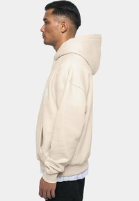 Beige Kapuzenpullover mit lockerer Passform, weichem Material und Fronttasche; über einem weißen Shirt getragen, mit gerippten Bündchen und Saum.