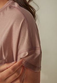 Camicia rosa chiaro a maniche corte realizzata in tessuto liscio, caratterizzata da cuciture piatte e una vestibilità morbida. Mano che tocca il bordo della manica.