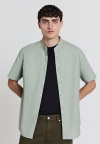 Hellgrünes kurzärmeliges Shirt mit Stehkragen, Knopfverschluss und weichem Material, über einem schwarzen T-Shirt getragen. Olivgrüne Hose.