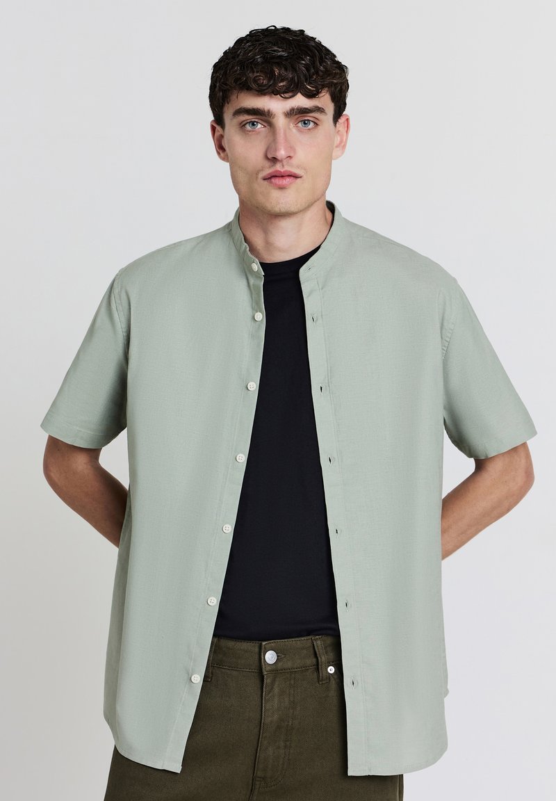 Hellgrünes kurzärmeliges Shirt mit Stehkragen, Knopfverschluss und weichem Material, über einem schwarzen T-Shirt getragen. Olivgrüne Hose.