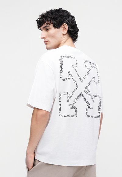 Νεαρός με σγουρά μαύρα μαλλιά, που φοράει λευκό φαρδύ t-shirt με αφηρημένο μαύρο σχέδιο κειμένου στην πλάτη, μπεζ παντελόνι, κοιτάζοντας πλάγια.