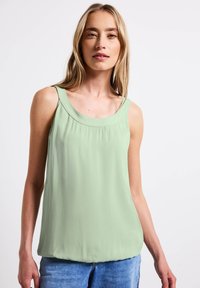 Světle zelený top bez rukávů s kulatým výstřihem, sklady na přední straně a příjemným, hladkým materiálem. V kombinaci s modrými džínovými šortkami.