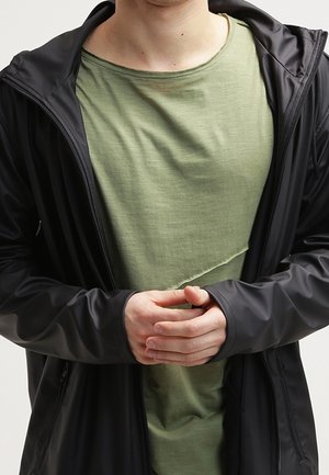 Veste noire imperméable avec capuche sur un t-shirt léger vert clair à col rond. La veste présente une texture lisse et des poignets ajustés, les mains croisées.