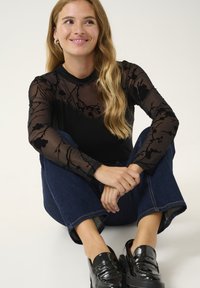 Černý top s dlouhým rukávem z průhledné látky s květinovým vzorem, spárovaný s tmavě modrými širokými džínami a lesklými černými mokasíny.