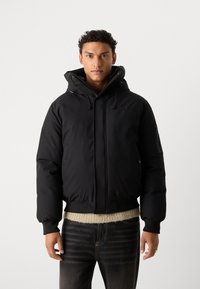 Black hooded puffer jacket with a zippered pocket, ribbed cuffs, and waistband. Smooth fabric with a matte finish. Fleece visible beneath hem.