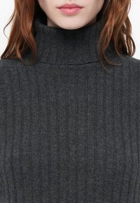 Ženska v temnosivi rebrasti pulover s turtleneck, ki prikazuje dekolte, ustnice in del valovite rdečkaste lase pred enobarvno ozadje.