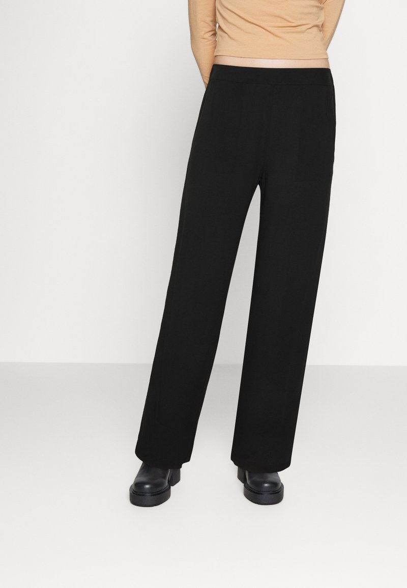 Personne portant un pantalon noir à jambes larges, des chaussures compensées noires, et un haut beige à manches longues, debout devant un fond blanc uni.