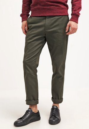 Homme portant un pantalon retroussé vert olive, des baskets noires et un haut à manches longues rouge, se tenant devant un fond blanc.