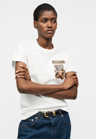 Γυναίκα με σταυρωμένα χέρια φοράει λευκό T-shirt Moschino με σχέδιο αρκουδάκι και σκούρο τζιν με μαύρη ζώνη.