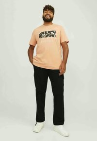 Tricou portocaliu cu mâneci scurte și model grafic negru, asortat cu pantaloni negri și adidași albi. Modelul stă pe un fundal neutru.