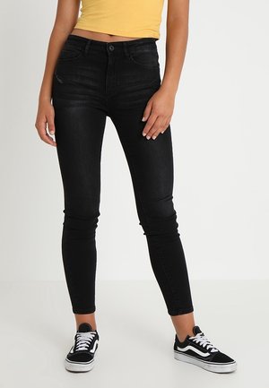 Femme portant un jean skinny noir, des baskets noires et blanches, et un débardeur côtelé court jaune, debout sur fond blanc.