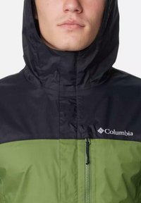 Grüne und schwarze Columbia-Jacke mit Reißverschluss, Kapuze und strukturiertem Stoff. Mit Logo auf der Brust und glatter Oberfläche.