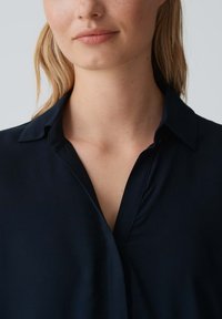 Blusa blu navy con colletto, caratterizzata da una texture liscia e una parte anteriore parzialmente aperta. Il tessuto sembra leggero e si drappeggia dolcemente.