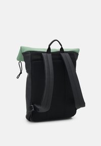 Ryggsäcken har en rulltoppsdesign med en mintgrön ovandel, svart nätmaterial, vadderade axelremmar och ett handtag för att bära den ovanpå.