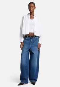 Camicia bianca corta con bottoni sopra una canotta bianca, abbinata a jeans larghi blu e ballerine nere a punta. Insieme semplice e casual.
