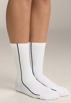 Nohy v bielych rebrovaných ponožkách s tenkým vertikálnym námorníckym pásikom od členka po lýtko na neutrálnom pozadí.