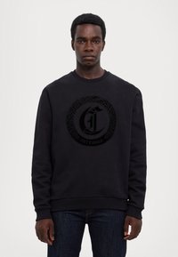 Felpa nera in cotone, con un logo circolare testurizzato al centro. Collo e polsini a coste, abbinati a jeans scuri.