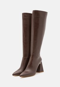 Paire de bottes en cuir marron, hautes avec des bouts pointus et des talons carrés, placées côte à côte sur un fond blanc.