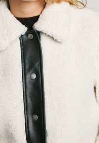 Fleece jacket featuring a white texture with a black leather front panel and snap buttons. Includes a collared neckline.