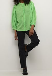 Green polka dot blouse with balloon sleeves, button closure, paired with black straight-leg pants and black shoes, against a neutral background.
