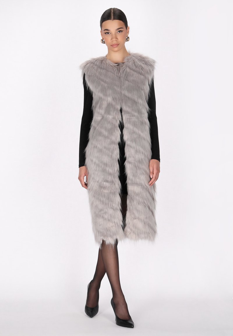 Chaleco largo de pelaje sintético gris con cuello, que presenta un corte recto, textura suave y apertura frontal. Combinado con una blusa negra y tacones.
