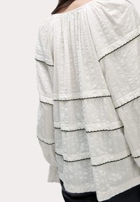 Blusa blanca con textura que presenta volantes en capas y ribetes negros, con un ajuste holgado y un escote fruncido. Tejido suave, ligeramente transparente.