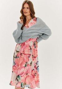 Kvetinové šaty s veľkými ružovými kvetmi a zelenými akcentmi, v kombinácii so svetlomodrým pleteným šálom, s uvoľneným, splývavým dizajnom.