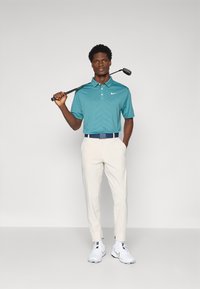 Πολυφορεμένο polo μπλουζάκι σε τιρκουάζ σχέδιο με κοντά μανίκια, κρεμ παντελόνι με κωνικό κόψιμο, ναυτικό ζώνη και λευκά sneakers. Το μοντέλο κρατάει ένα golf club πάνω από τον ώμο του.