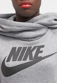 Grå hoodie tillverkad av mjukt material med en stor svart "Nike"-logotyp och en stiliserad swoosh-emblem. Huvan har en bekväm, lös passform.