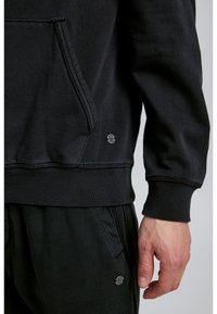 Melna kapuce ar priekšējo kabatu, ribinātām aprocēm un apakšmalu. Uz sāna ir neliels logo elements. Mīksts, teksturēts audums.