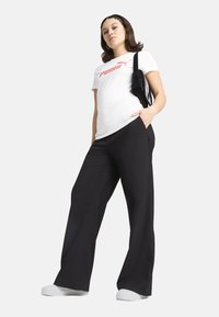 T-shirt bianca con logo "PUMA" rosso, abbinata a pantaloni neri a gamba larga e sneakers bianche. Il modello tiene una borsa nera. Outfit semplice e casual.