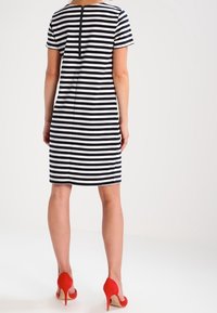 Kvinna klädd i en svart- och vitrandig knälång klänning med korta ärmar och röda högklackade skor, stående mot en enkel bakgrund.