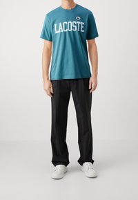 Tyrkysové bavlněné tričko s bílým nápisem "LACOSTE" a logem, v kombinaci s černými kalhotami a bílými teniskami, minimalistický design.