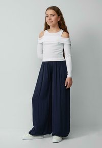 Weißes, schulterfreies Oberteil mit langen Ärmeln und geripptem Muster, kombiniert mit navyblauen, weiten, gefalteten Hosen und weißen Sneakern.