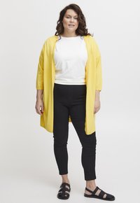 Žlutý lehký kardigan přes bílý tričko, doplněný černými slim-fit kalhotami a černými sandály. Kardigan má 3/4 rukávy a volný střih.
