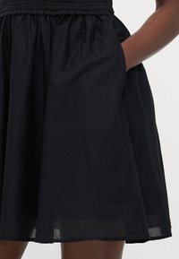 Vestido negro con un cuerpo fruncido, falda acampanada, bolsillos laterales y una tela ligera y texturizada. El dobladillo cae justo por encima de la rodilla.