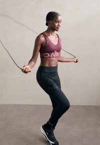 Braidelt hajjal rendelkező nő bordó sportmelltartót és fekete leggingset visel, és ugrál a kötéllel egy minimalista belső térben.