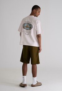 Light pink cotton t-shirt with a graphic design on the back; paired with olive green knee-length shorts and light-colored slip-on shoes.